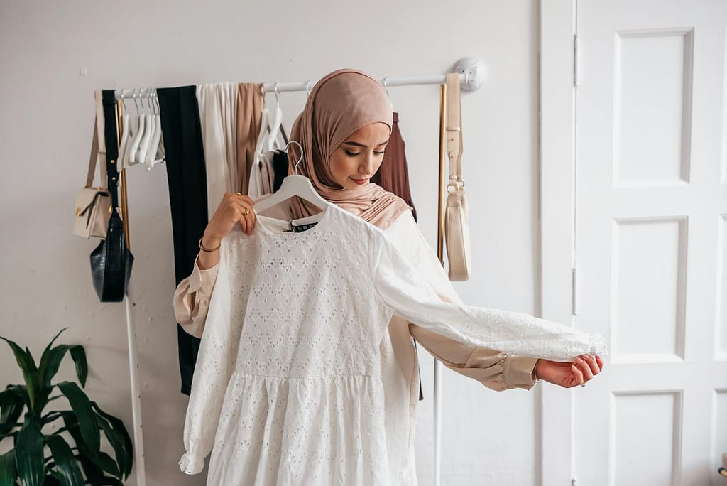 A Muslim woman in a hijab examines a white dress in a clothing boutique with racks of clothes.