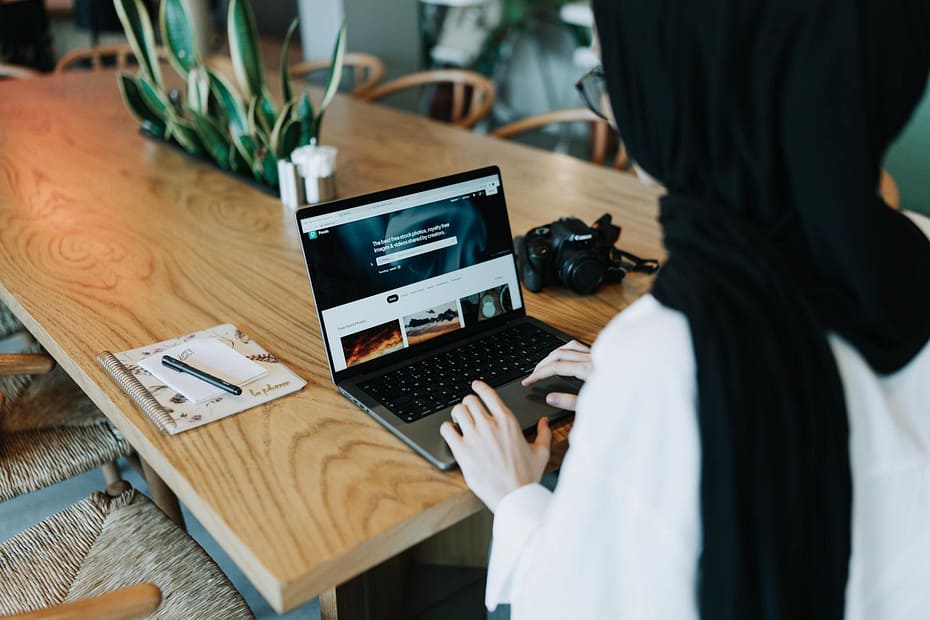 Woman in hijab using laptop at wooden table in modern office with digital camera and plants.
