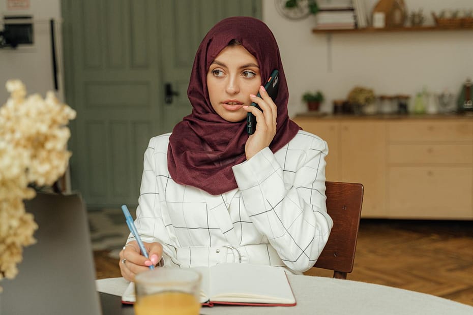 Young woman in hijab using smartphone and writing notes at home office, reflecting modern technology and remote work.