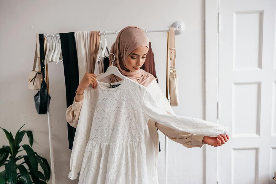 A Muslim woman in a hijab examines a white dress in a clothing boutique with racks of clothes.